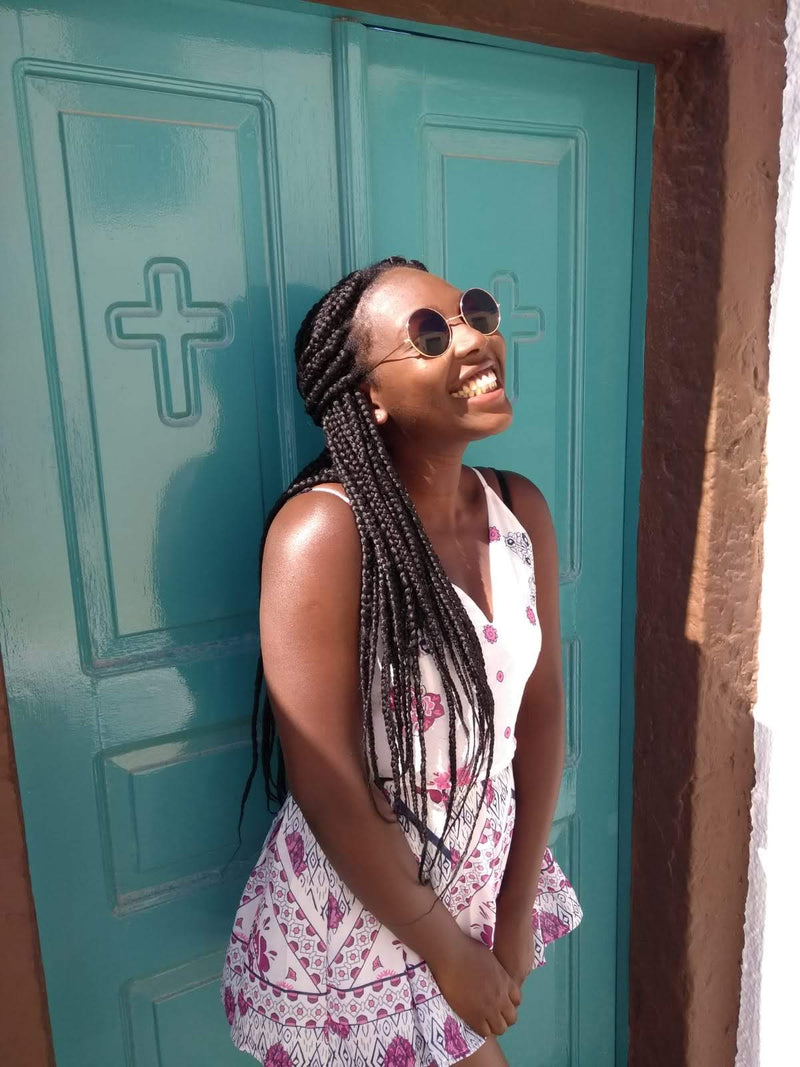 Woman with braided hair wearing sunglasses and a patterned dress standing in front of a teal door.